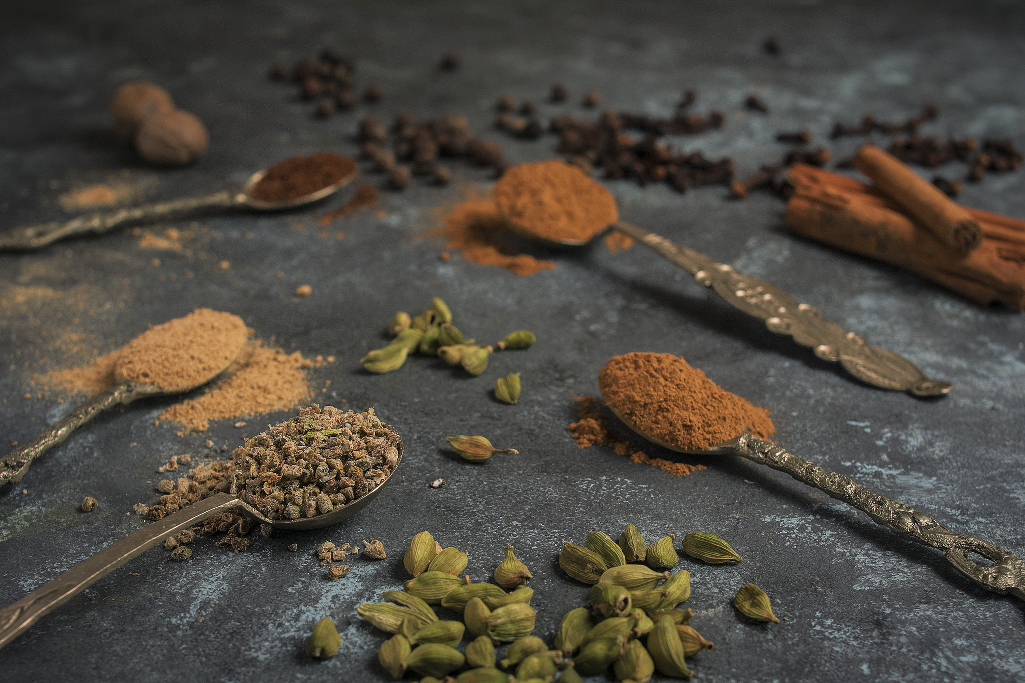 Ground spices on vintage spoons with cardamom pods, cinnamon sticks, cloves and nutmeg on a dark surface.
